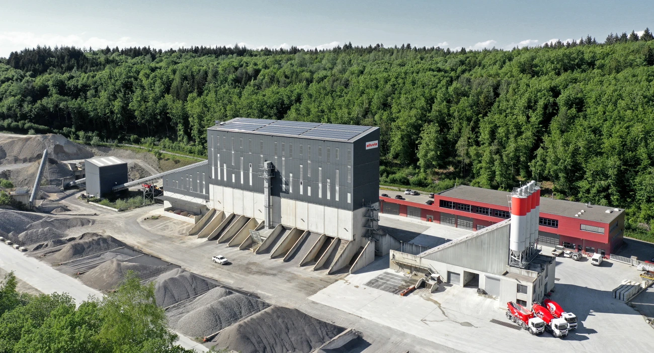 Kies- und Betonwerk Oberwangen mit rotem Betriebsgebäude und Wald im Hintergrund. Im Vordergrund drei rote LKWs der alluvia Flotte.