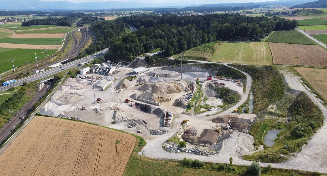 Luftaufnahme des Kieswerks Mattstetten umgeben von landwirtschaftlichen Feldern und einem dichten Waldgebiet. Die Grube enthält mehrere Bagger und schwere Maschinen, die in verschiedenen Bereichen des Geländes arbeiten. Neben der Anlage verläuft eine Bahnstrecke und eine Autobahn mit vorbeifahrenden Fahrzeugen.