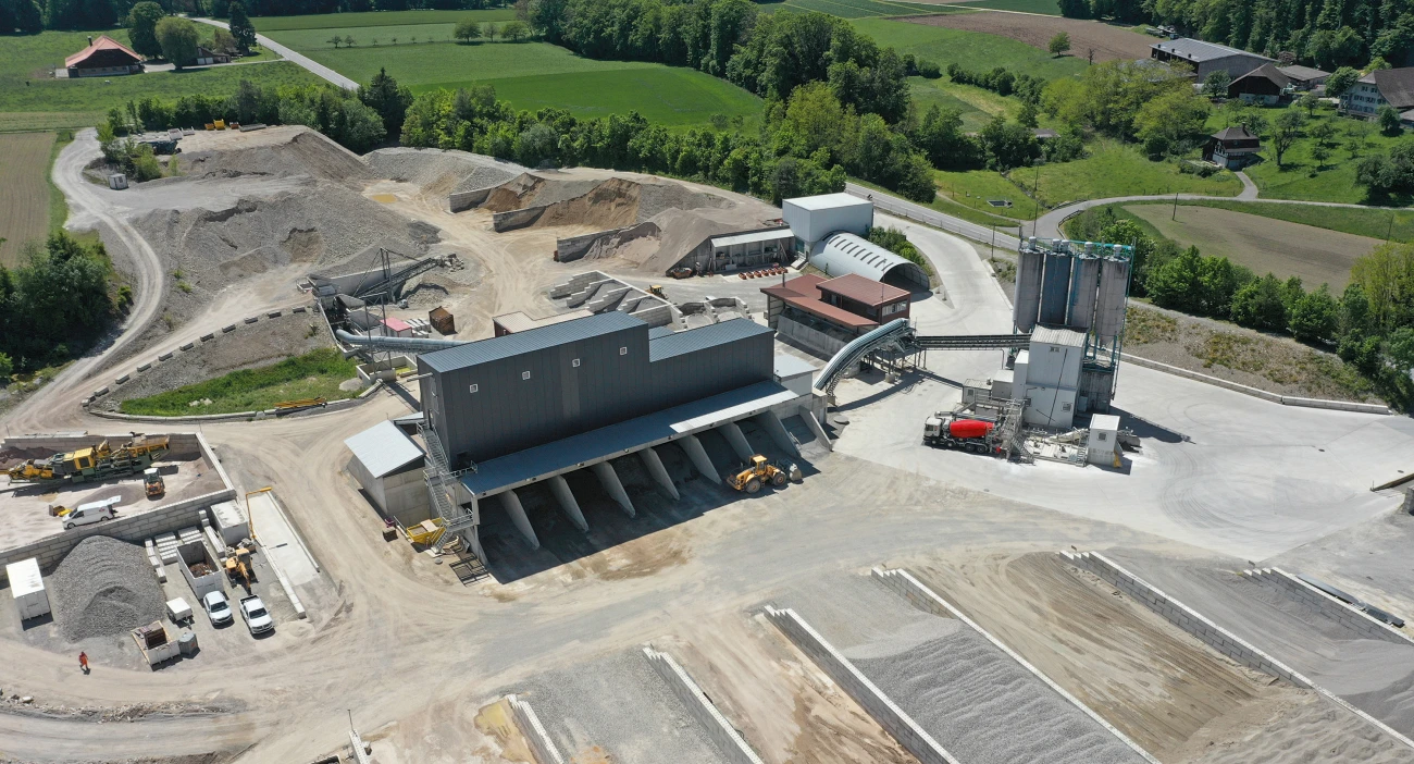 Drohnenansicht vom Kies- und Betonwerk in Berken. Im Zentrum das graue Kieswerk, im Vordergrund verschiedene Kiessilos und im Hintergrund Wiesen und Wald der Gemeinde Berken.